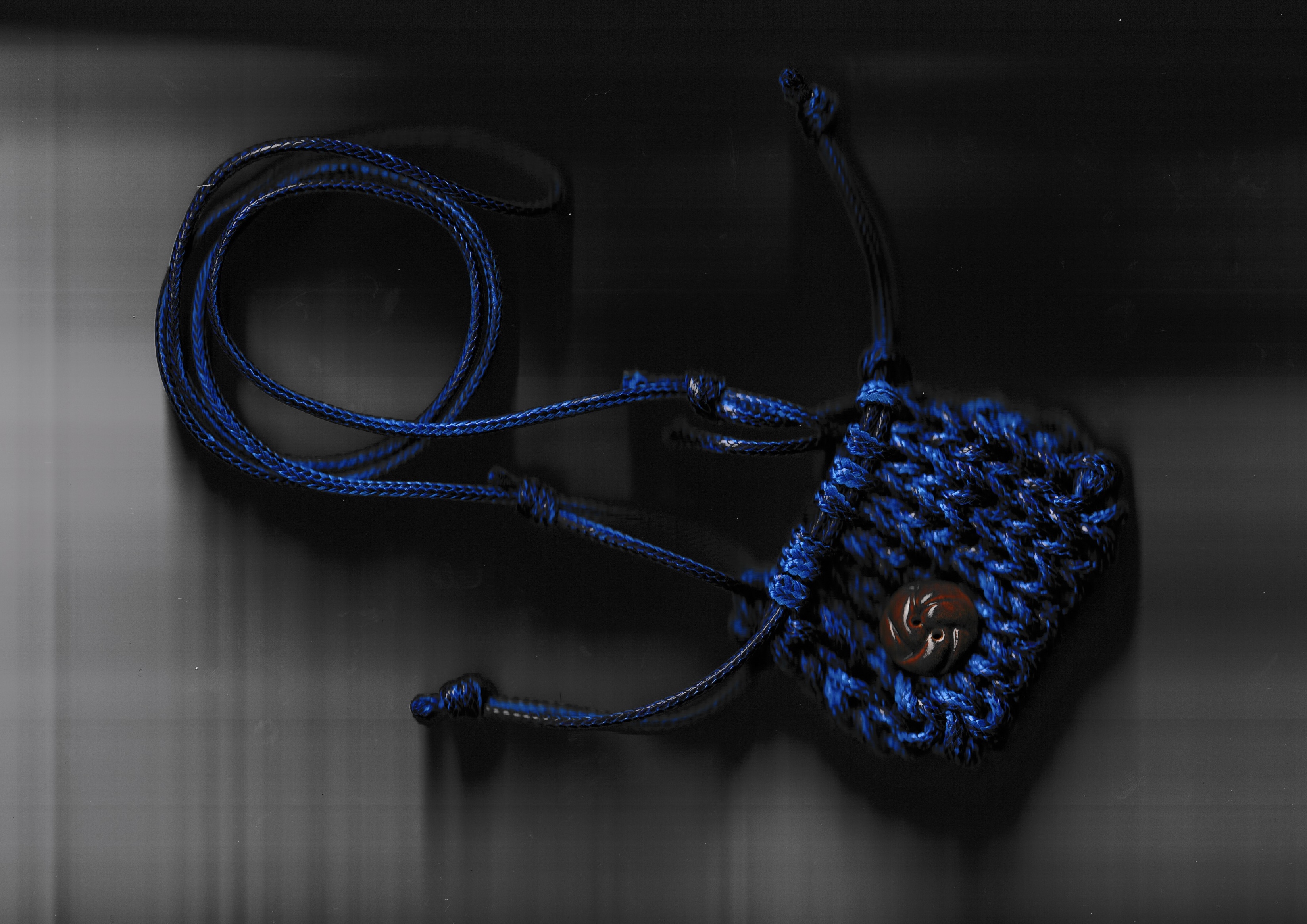 A small hand-knitted blue drawstring pouch on a cord, with a carved bead, photographed on a dark reflective surface.