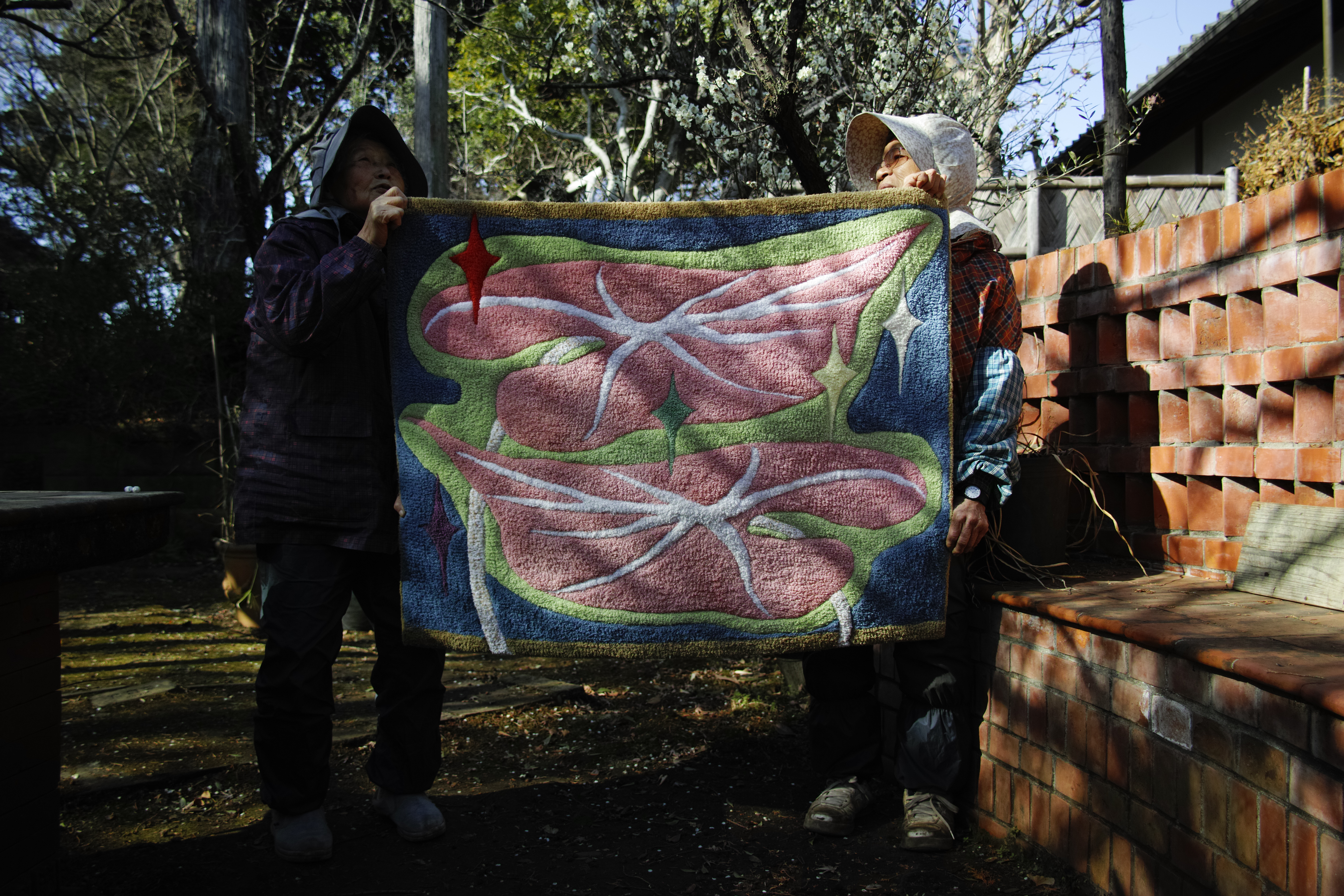 Two people holding a large hand-tufted rug outdoors at Daichuji temple, its pink and blue forms facing the camera.